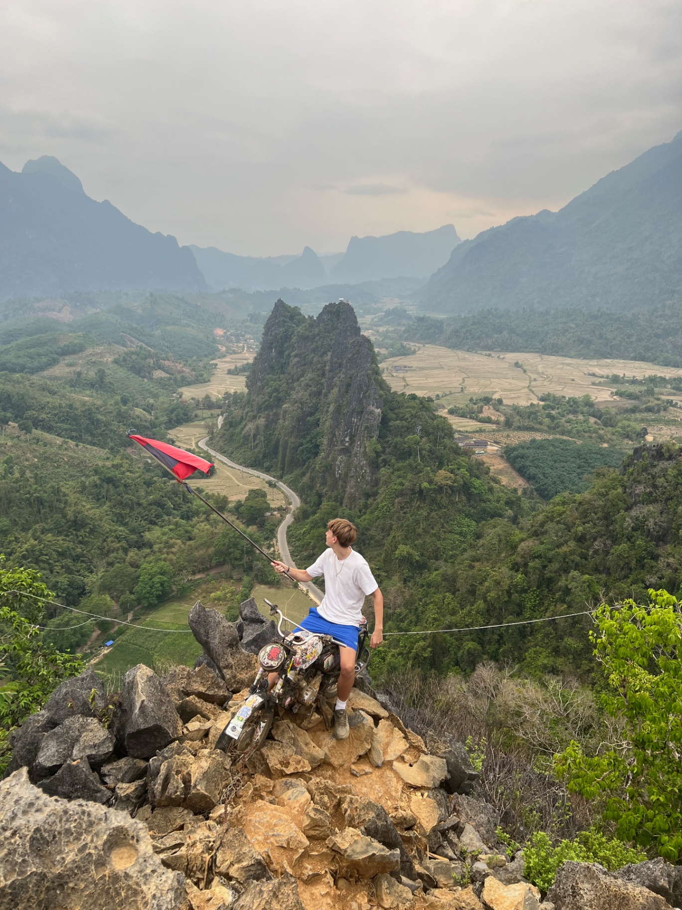 Vang Vieng — Flag in hand on a limestone peak above the karst valley and rice paddies.