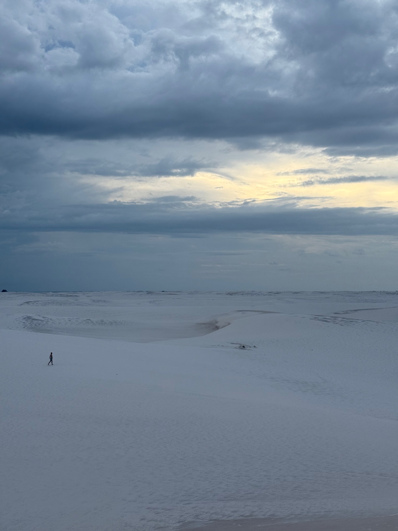 Lencois Maranhenses — A lone walker crossing the pale dunes under heavy sky.
